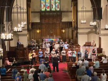 A large choir sings before a congregation in the front of a gothic revival church with stained glass windows, a large pipe organ, and hanging chandeliers.