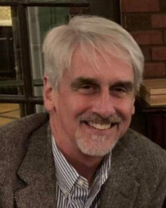 Gordon Moriarty, a man with gray hair and beard, wearing a striped button-down shirt and tweed jacket.