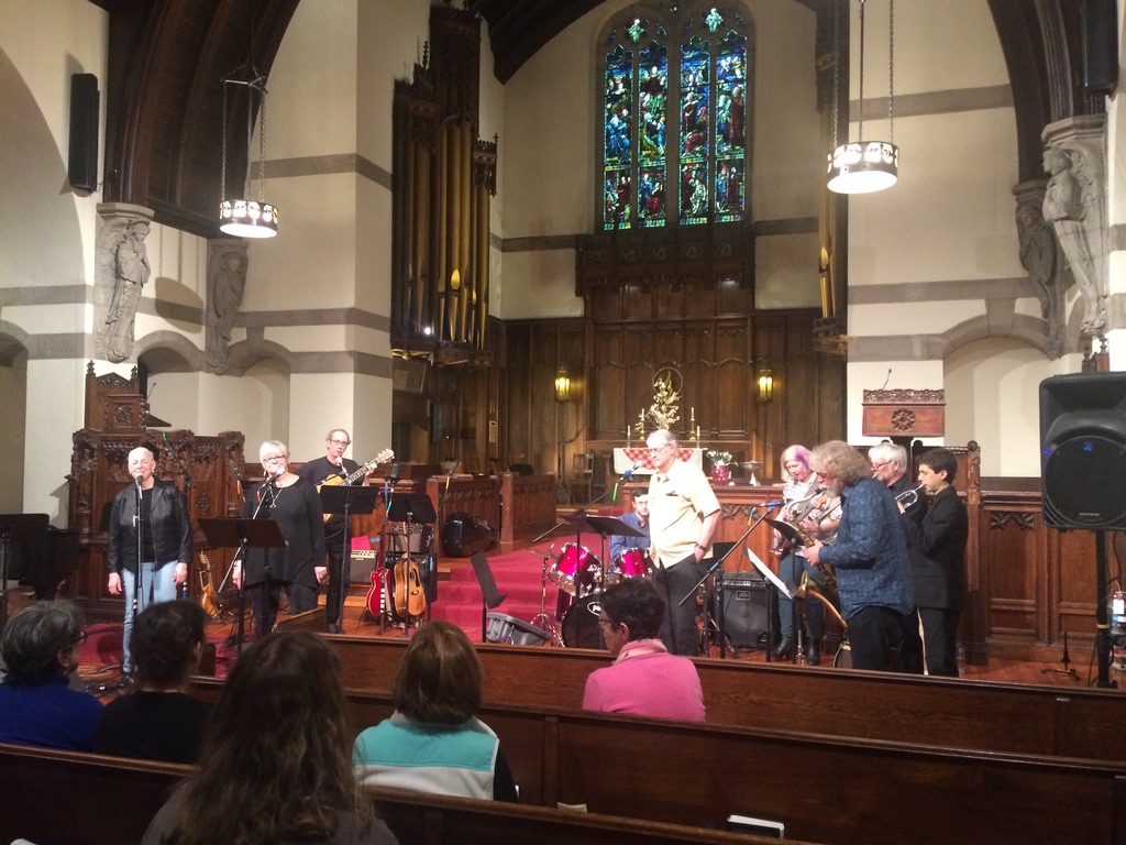 FUUSN Band - A group of adult sing and play instruments including guitars, drums, trumpets, and trombones before a congregation in a church.