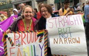 Two women at a parade, one in a pink shirt and black jacket holding a sign with a rainbow border reading, "PROUD FAMILY", the other wearing glassed and a black coat, holding a sign reading, "STRAIGHT BUT NOT NARROW."