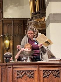 In a church with a pipe organ in the background, a woman with shoulder-length brown hair wearing a purple shirt and gray sweater assists a red-haired boy as he lights a candle inside a large silver chalice.