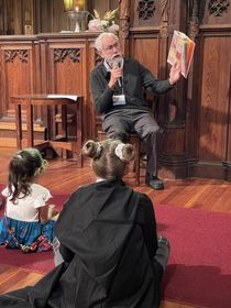 A man with white hair, mustache, and beard wearing glasses, a dark gray sweater over a blue button-down shirt and gray pants reads from a book to two children sitting on a red rug over a wood floor.