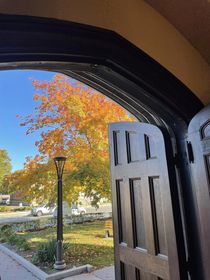 Large wooden doors in an arched doorway open to the church front yard, with a sidewalk, hedge, lawn, lamppost, and maple tree in fall colors ranging from green to yellow to bright orange.