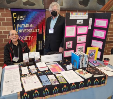 Two adults run an outreach table for the Racial Justice Ministry, including a quilted banner reading "First Unitarian Universalist Society of Newton," a posterboard, a selection of books, and many pamphlets and flyers.