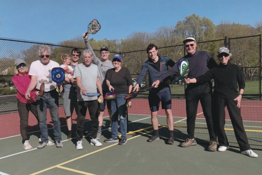 Nine adults in activewear, one holding a small child, stand on a tennis court with racquets and balls.