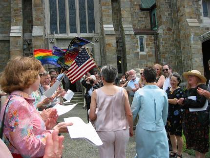 two women walk arm in arm with their backs to the viewer through a crowd of people cheering and waving rainbow and US flags