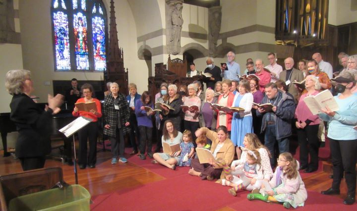 A large group of adults and children, standing and seated, perform in the sanctuary of a church with stained glass windows in the background.