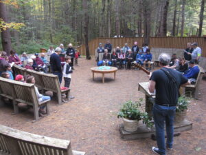 A worship service with about thirty people of all ages is held in an open-air chapel among tall pines. In the foreground, a man stands and plays a guitar before a podium.