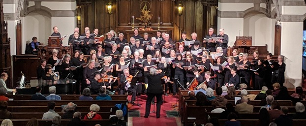 A large choir of adults dressed in black stand in the sanctuary of a gothic revival church and sing to a congregation, accompanied by stringed instruments and piano, and conducted by a gray-haired woman in a formal black pantsuit.