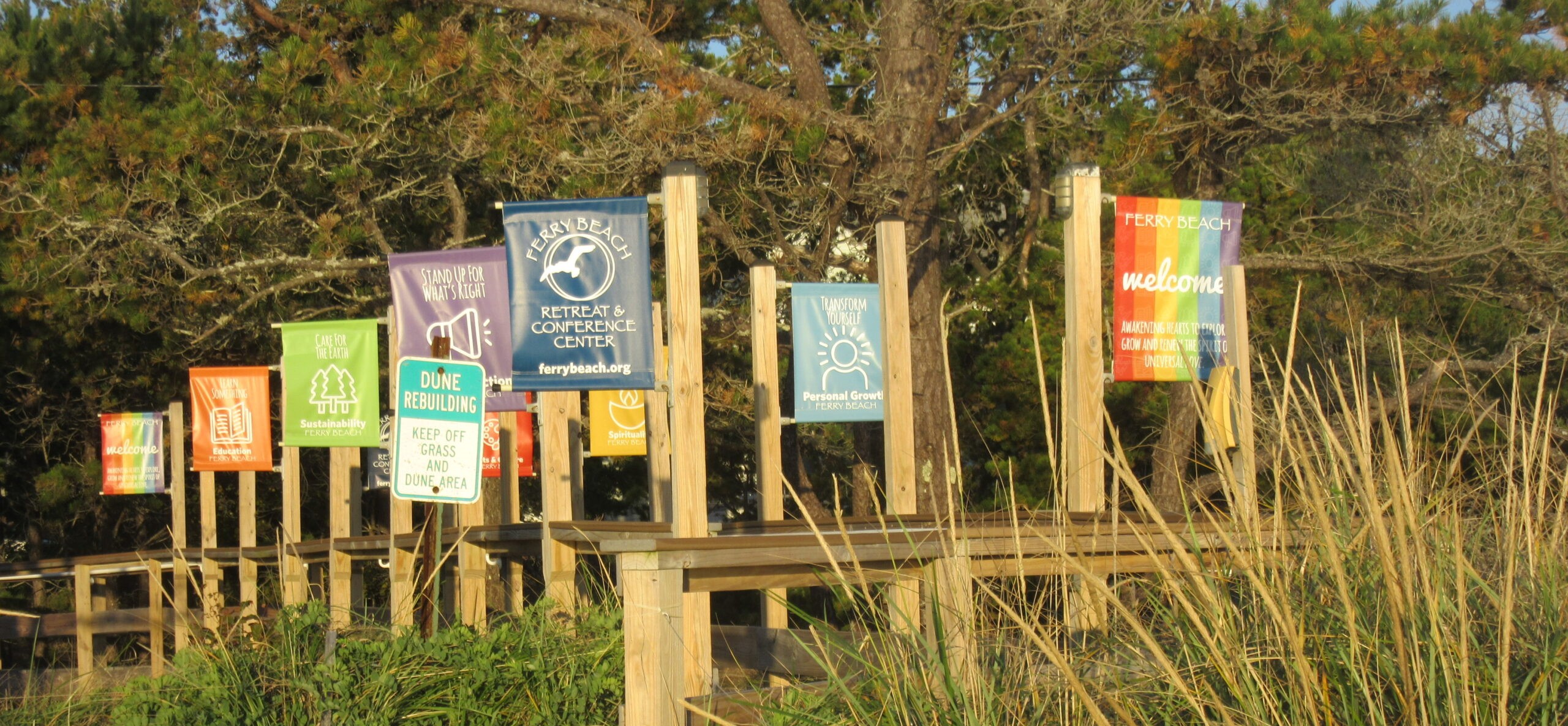 Many multicolored flags on wooden poles wave along a beach with grass and shrubs. A blue flag reads, "Ferry Beach Retreat and Conference Center - ferrybeach.org." A rainbow flag reads, "Ferry Beach - WELCOME - Awakening Hearts to Explore, Grow, and Renew the Spirit of Universal Love." A purple flag reads, "Stand Up for What's Right."