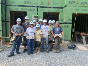 A group of ten adults wearinig hard hats stand before a home under construction.