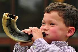 a smiling, round-faced boy blows a ram's horn (Shofar)