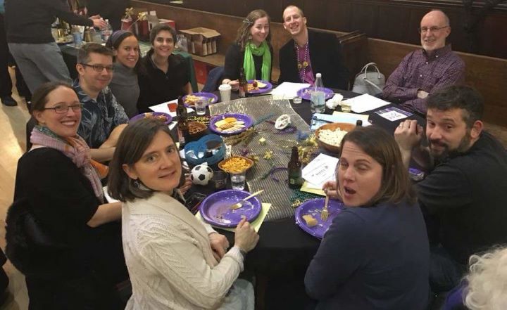 Ten adults seated at a round table with Mardi Gras-themed decorations eat snacks and smile.