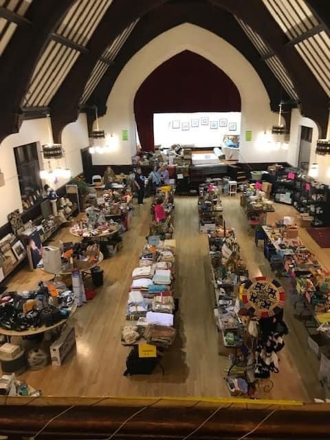 Viewed from above the Parish Hall, four rows of folding tables are covered in items for sale. Shelves line one wall of the hall and pews the opposit walls, laden with further selections. The stage at the far end has been largely cleared. All is ready for the sale.