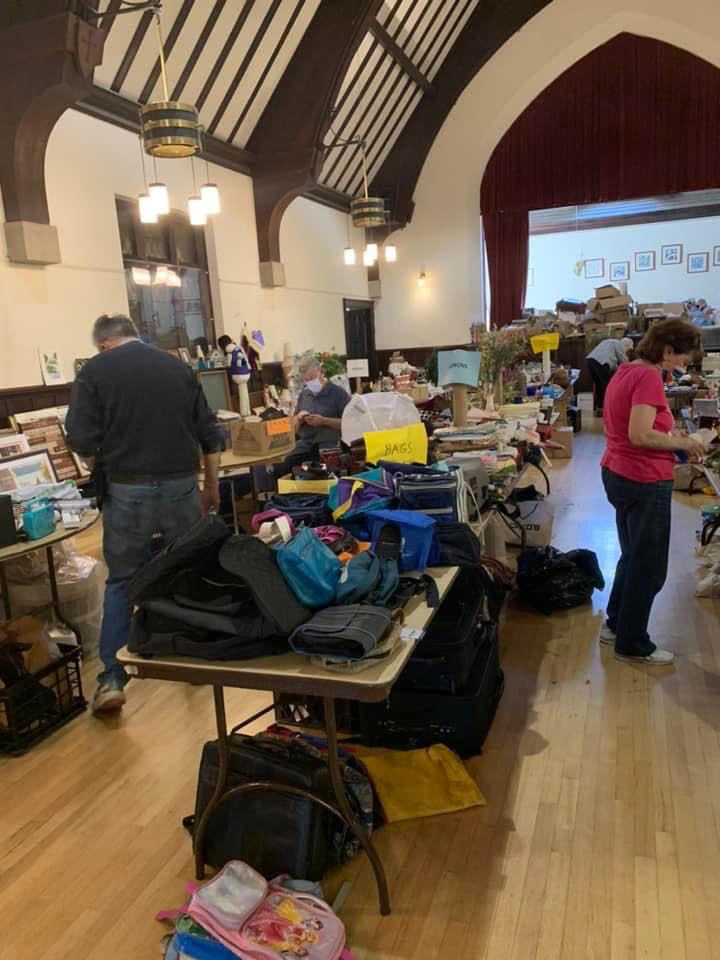 Volunteers arrange items such as bags and books on folding tables in the Parish Hall.