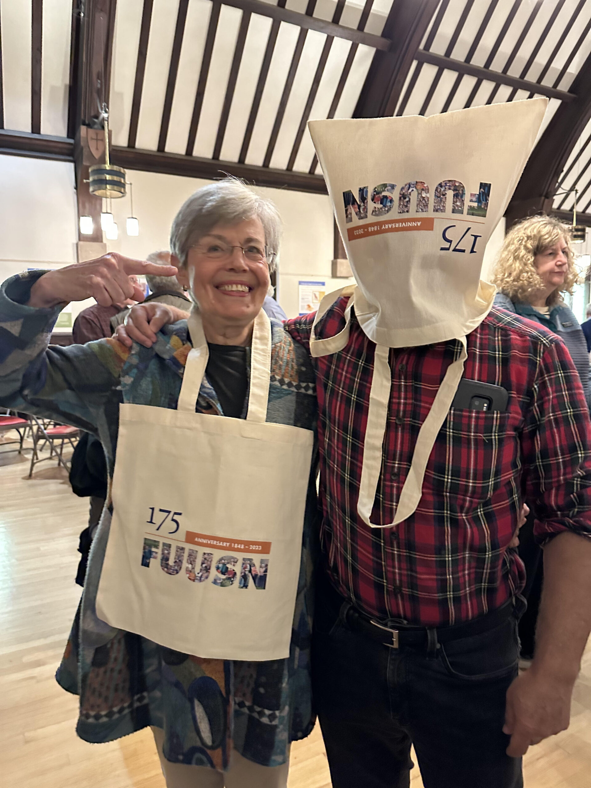 A man demonstrates a novel use for a 175th Anniversary tote bag by wearing it on his head. A woman next to him, wearing another tote around her neck, smiles and points at the man.