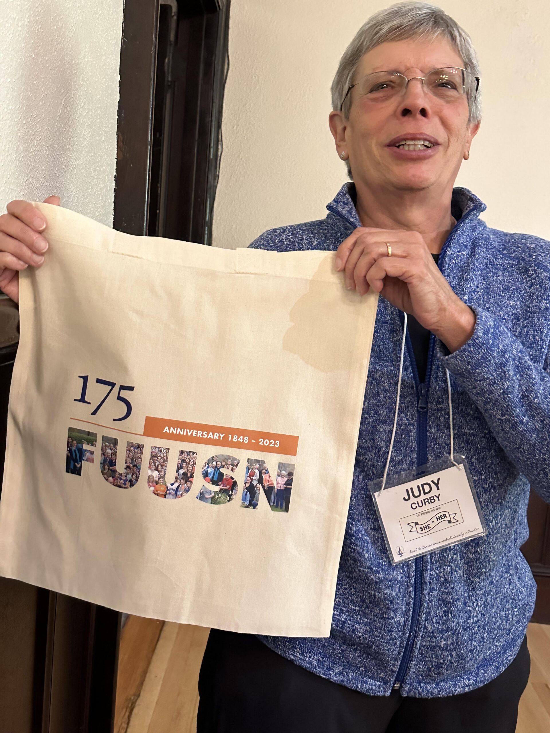 A woman with short gray hair and glasses in a blue zipped flannel sweater proudly holds up her 175th Anniversary tote bag.