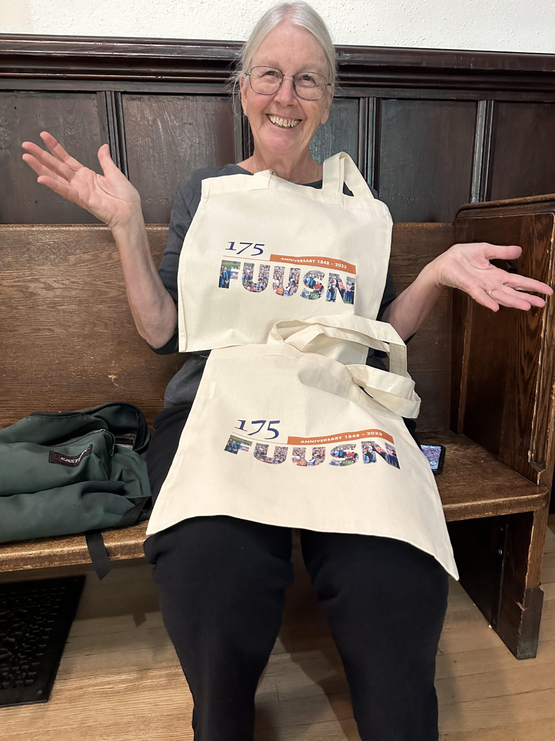 A woman with short gray hair and glasses seated on a church pew is blanketed with two 175th Anniversary tote bags.
