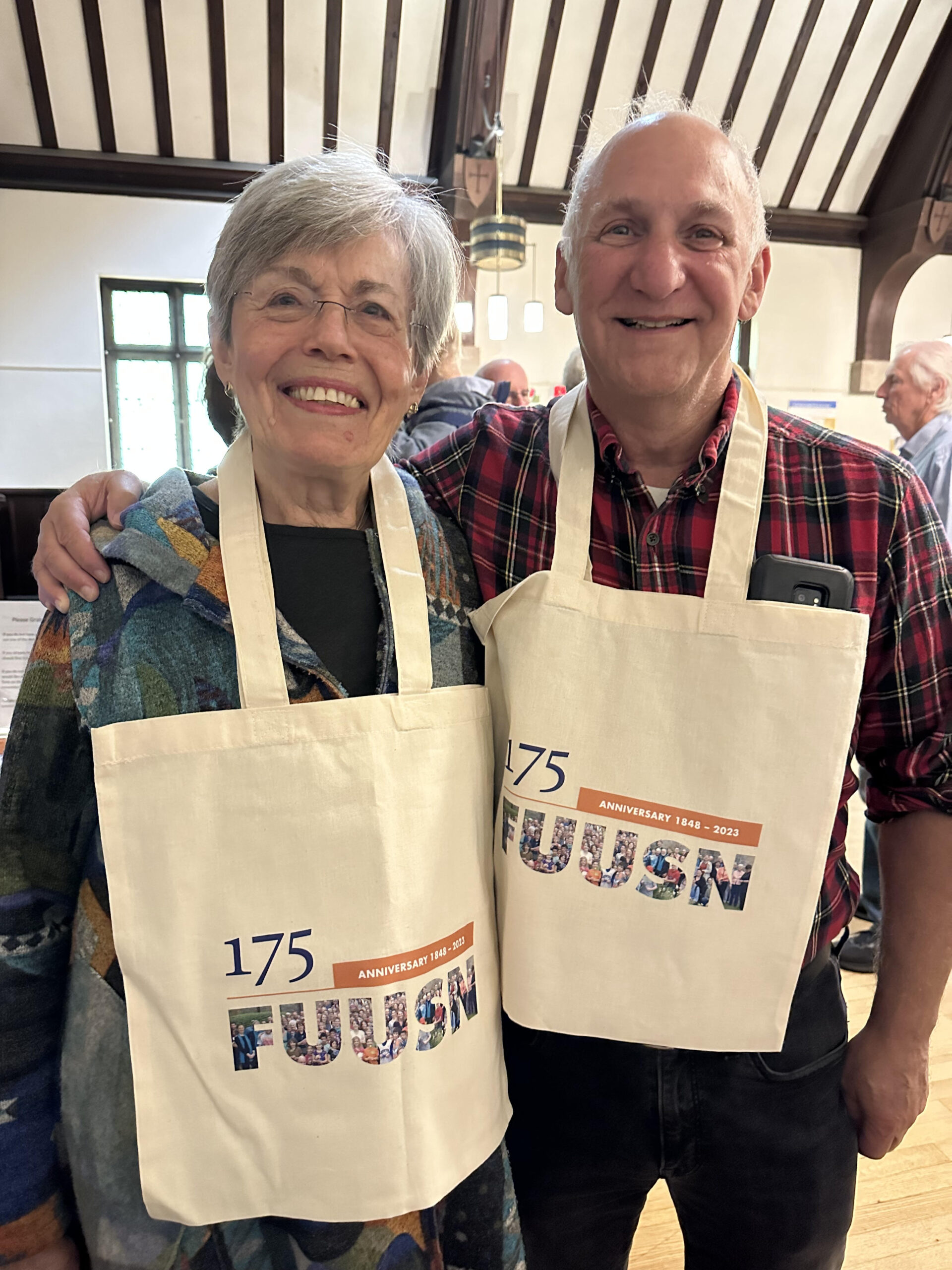 A man and a woman pose with 175th Anniversary tote bags hanging around their necks.
