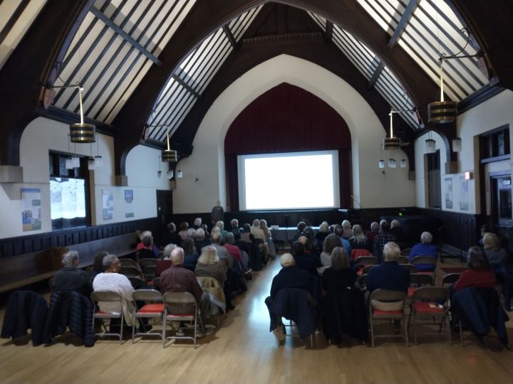 A large crowd of people sits on folding chairs in a darkened Parish Hall viewing a movie projected onto a screen.