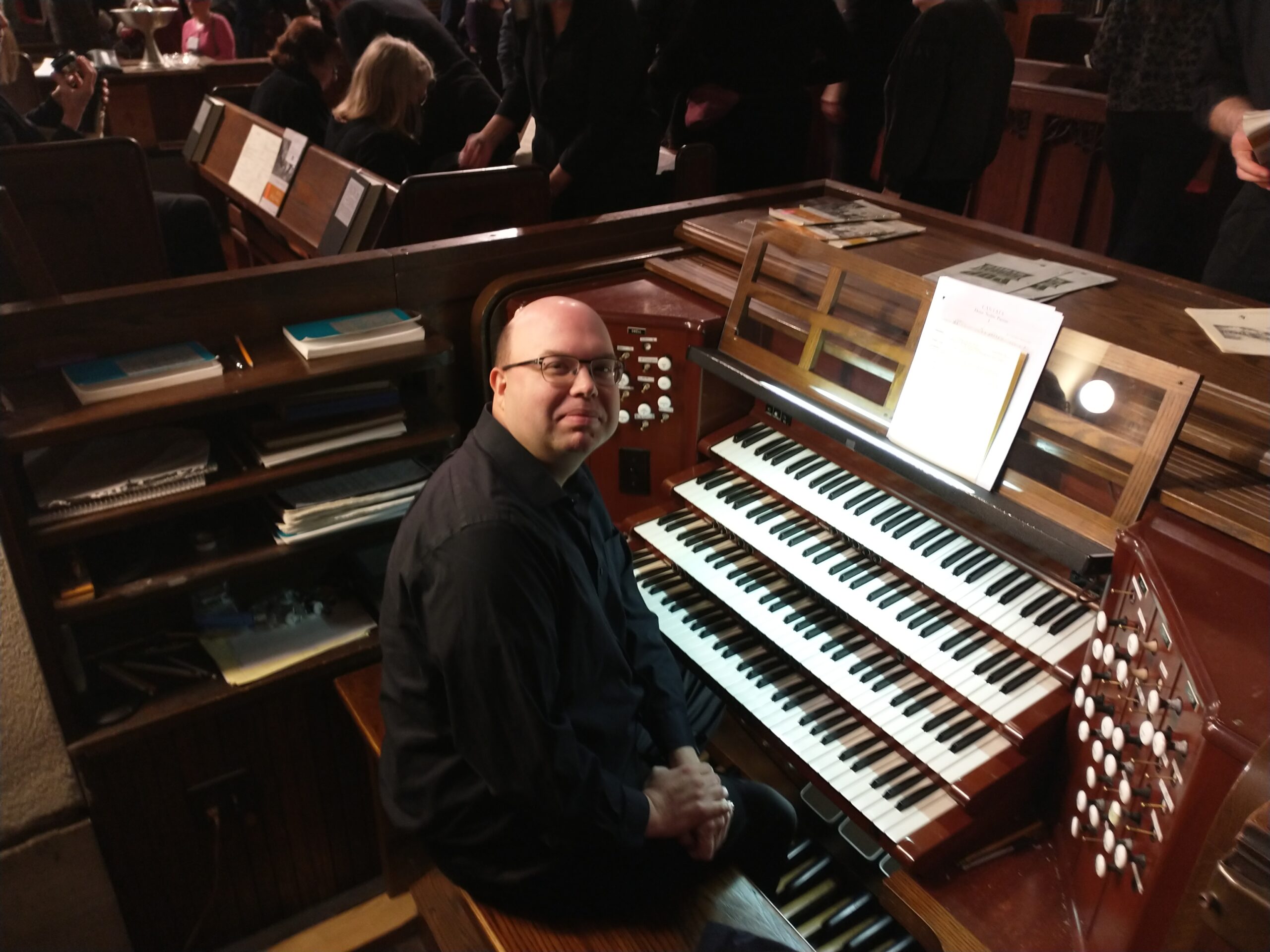 A bald man with glasses dressed in a black long-sleeved shirt and black pants sits at the complex keyboard of a pipe organ, with seated choir members in the background.