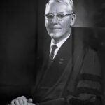 Black and white portrait of an older man with grey hair and glasses, seated in ministerial robes with his hands folded upon his lap.
