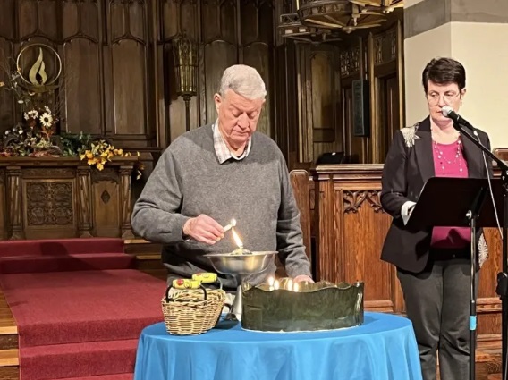 A gray-haired man in a gray sweater and white collared shirt lights a chalice in the front of the Sanctuary, while a dark-haired woman with glasses wearing a black jacket, pink shirt, and long necklaces stands behind a podium nearby.