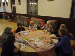 Several adults and children sit at a round table in the Parish Hall coloring and assembling crafts.