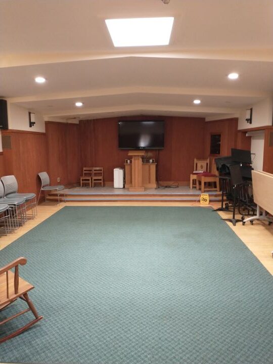 A small open chapel, with a podium, large-screen TV, skylights, and chairs, folding tables, and music stands lining the walls.