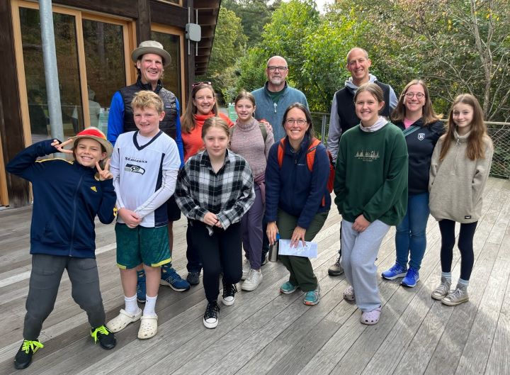 Six middle schoolers and five adults wearing sweaters and light jackets stand outside on a wooden deck at the visitors' center at Walden Pond, with green trees in the background