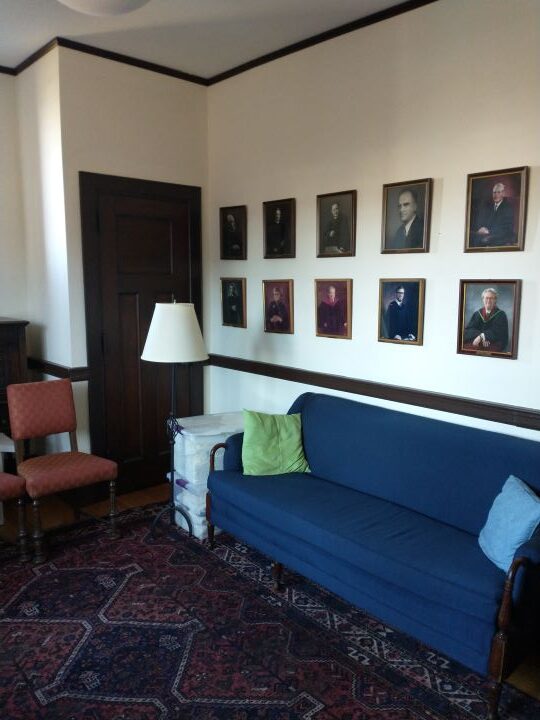 A cozy room with a large blue sofa and chairs and a floor lamp, with a double row of portraits hanging on the wall.