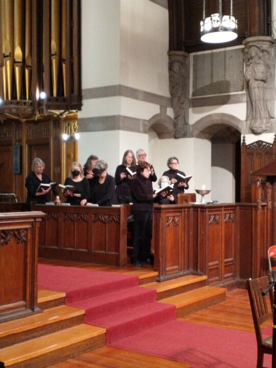A group of about ten adults wearing black stand in the choir seating in the Sanctuary and sing from hymnals.