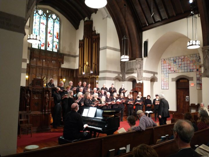 A large choir stands at the front of the Sanctuary and sings, accompanied by a male pianist and directed by a gray-haired woman dressed in black.