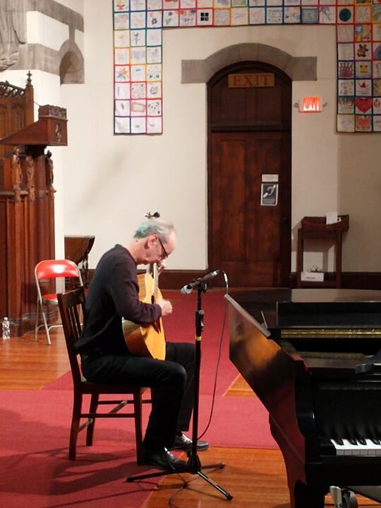 A man with gray hair wearing glasses, a black long-sleeved shirt, and black pants, sits in a chair beside a grand piano and plays the guitar.