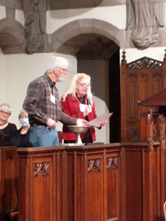 A man with white hair and beard wearing glasses, a plaid shirt, and blue jeans, lights the chalice as a woman with long, white hair wearing glasses and a red sweater reads from a sheet of paper.