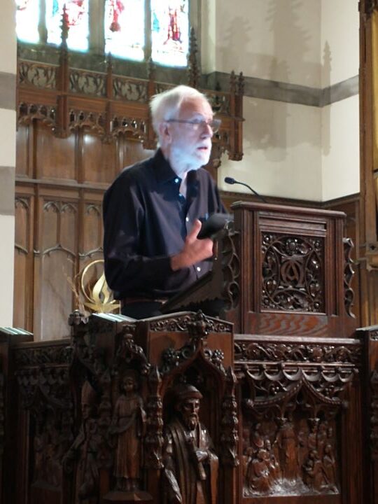A man with white hair and beard wearing glasses and a black long-sleeved shirt stands in the pulpit of the Sanctuary.