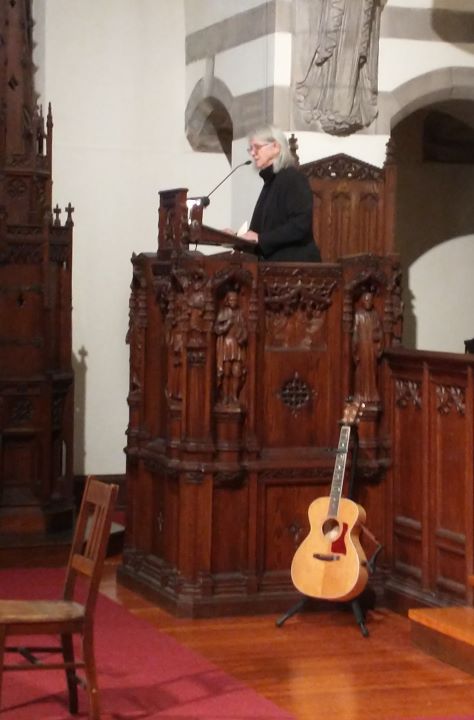 A gray-haired woman with glasses, dressed in a black suit and black shirt, stands in the pulpit. A guitar rests against its base.