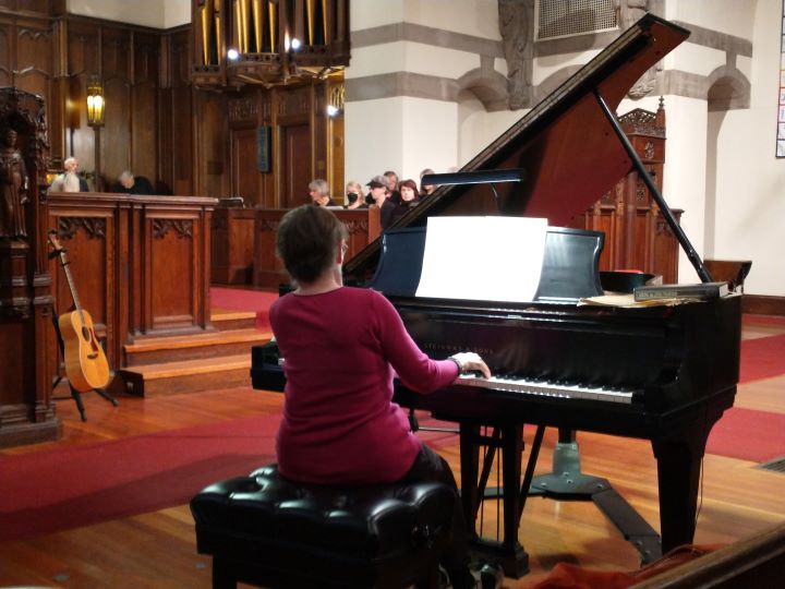 A woman with short brown hair wearing a long-sleeved pink shirt and black pants sits and plays a grand piano at the front of the Sanctuary