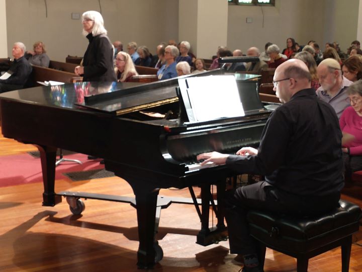 A bald man with glasses, dressed in a black long-sleeved shirt and black pants, sits and plays a grand piano in the Sanctuary, with a gray-haired woman standing nearby and many congregants seated in pews.