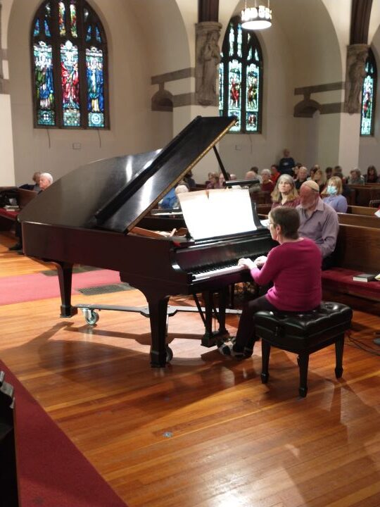 A woman with short brown hair wearing glasses, a long-sleeved pink shirt, and black pants sits and plays a grand piano while congregants in the pews listen.
