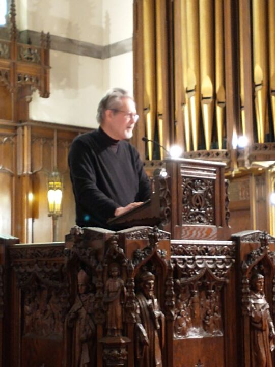 A man with gray hair and a white beard wearing glasses and a long-sleeved black shirt stands in the pulpit, with the pipe organ in the background.