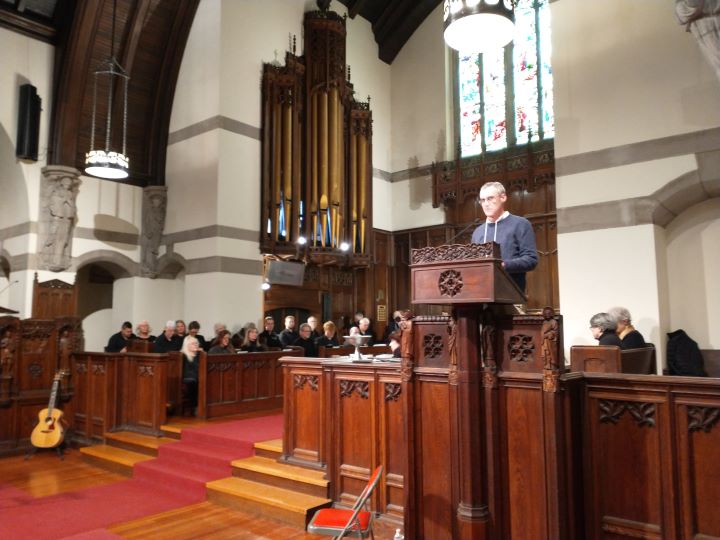 A man with gray hair and glasses wearing a gray sweater stands at a podium at the front of the Sanctuary, with choir members seated behind him.