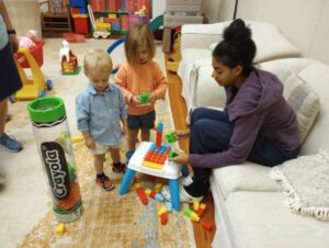 A teenager with black hair wearing a purple hoodie and blue jeans plays with brightly colored blocks with two toddlers, a blond boy in a blue shirt and tan shorts and a brown-haired girl in an orange shirt and blue shorts.