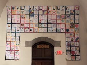 A quilted tapestry formed of many small squares forming an arch over the exit from the church Sanctuary. It reads "WE ARE FUUSN."