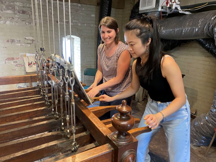 Two women press on wooden levers attached to steel cables which ring the chime (church bells)