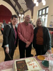 Rev. Parisa Parsa (a woman with short, black hair wearing glasses, a black jacket, and a pink shirt), Carson Cooman (a bald man wearing glasses and a red button-down shirt) and Anne Watson Born (a woman with shoulder-length gray hair wearing glasses, an orange turtleneck shirt, and a black sweater) stand before a table with a cut sheet cake as many other people talk and interact in the Parish Hall behind them.