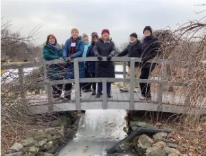 Seven adults dressed warmly in jackets, coats, and winter hats, stand on a bridge over a frozen stream.