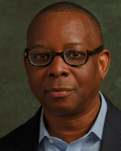 A Black man with rounded glasses and a serious expression, wearing a collared shirt and jacket