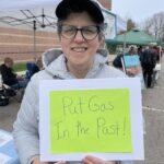 At an outdoor event, a woman with short brown hair wearing glasses, a baseball cap, and a gray hoodie holds up a sign on green paper that reads, "Put Gas In the Past!"