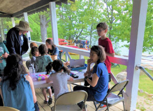Children and adults seated and standing at tables on a covered porch participate in crafts activities at Sandy Island.
